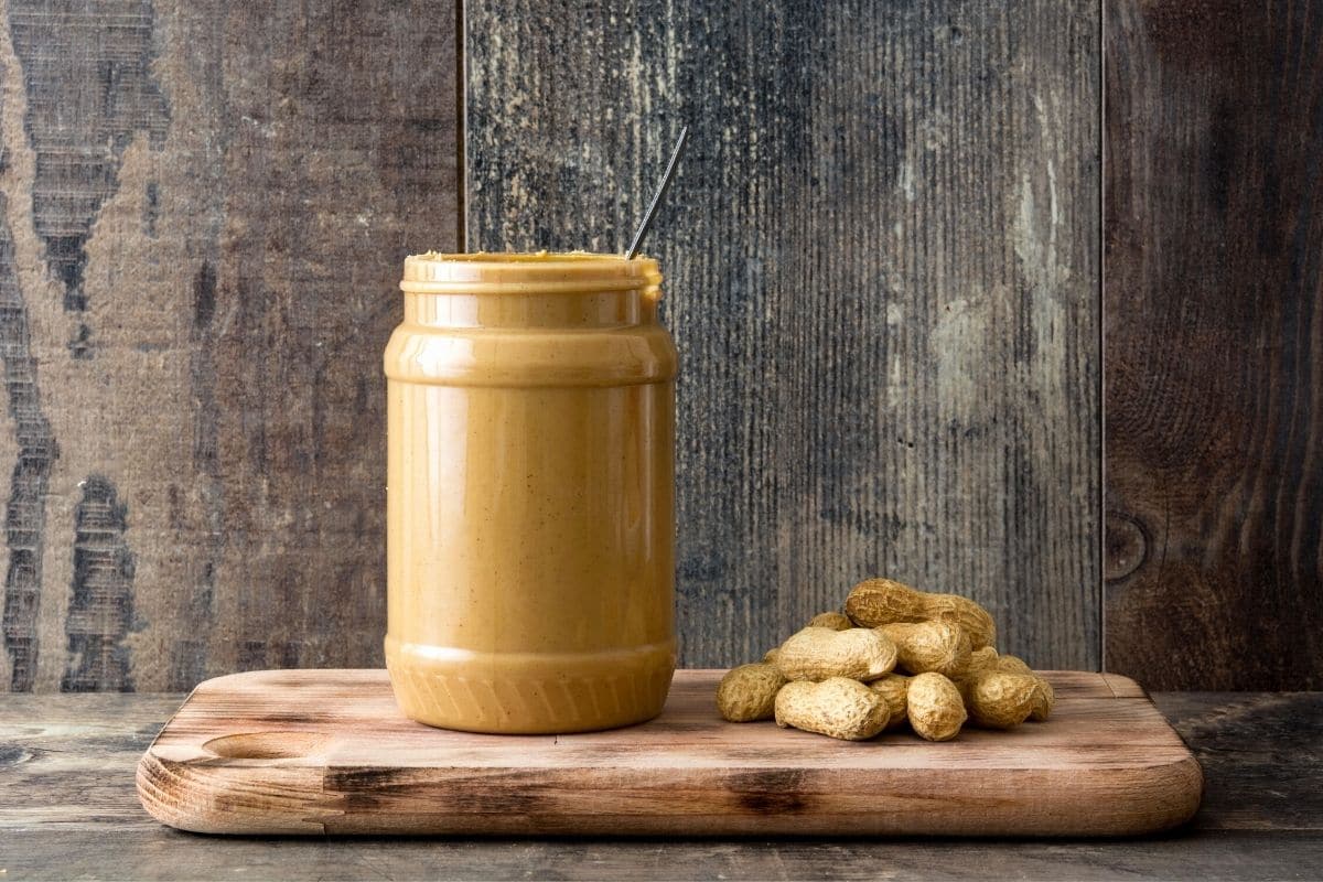 A jar of peanut butter on a wooden cutting board with peanuts next to it.