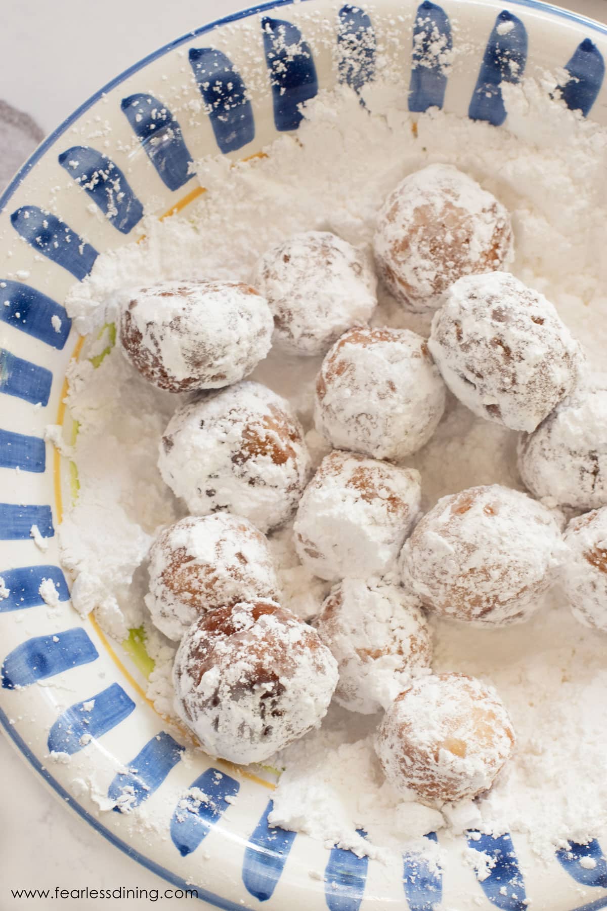 A bowl with powdered sugar and some donut holes getting coated in the sugar.