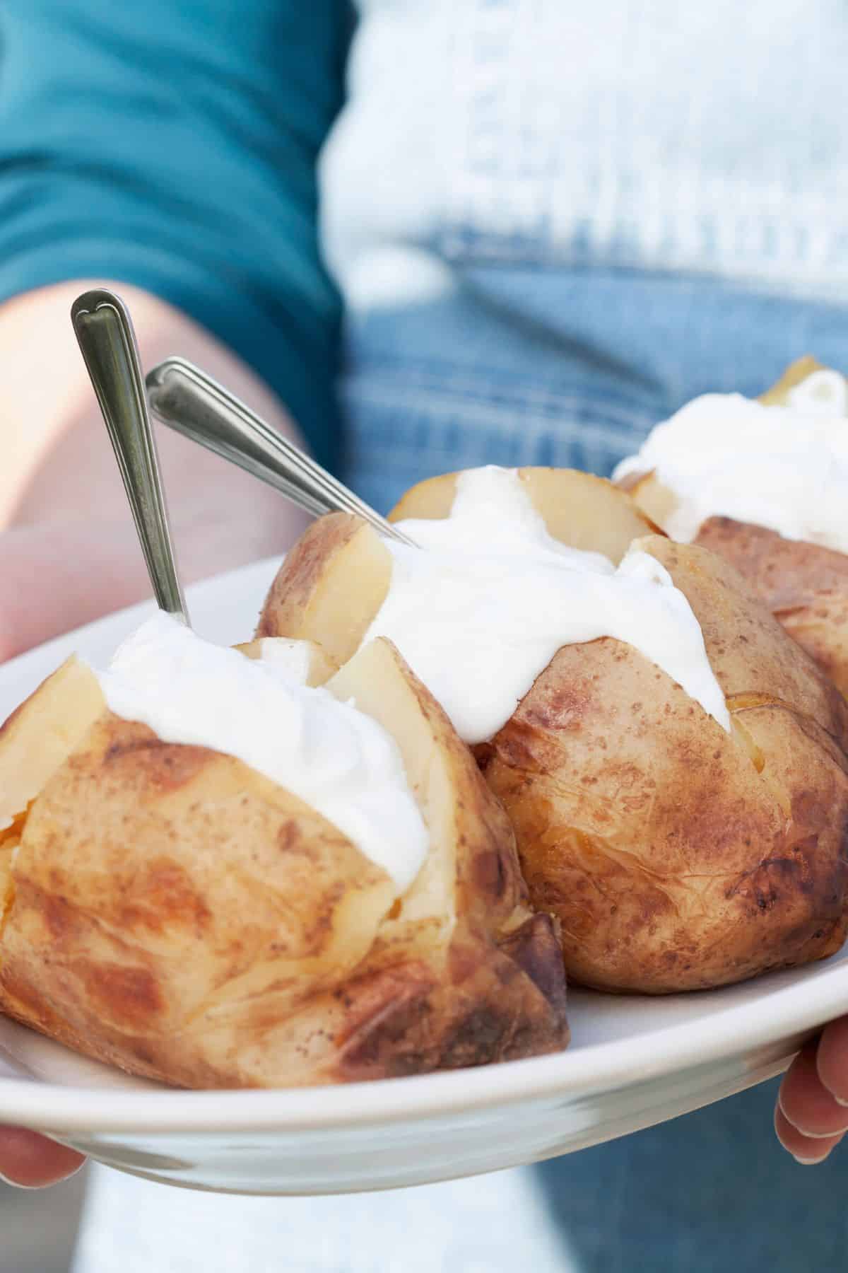 a person holding a plate with three baked potatoes topped with sour cream.